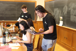 A group of students in a classroom, one is standing with their laptop advising another student